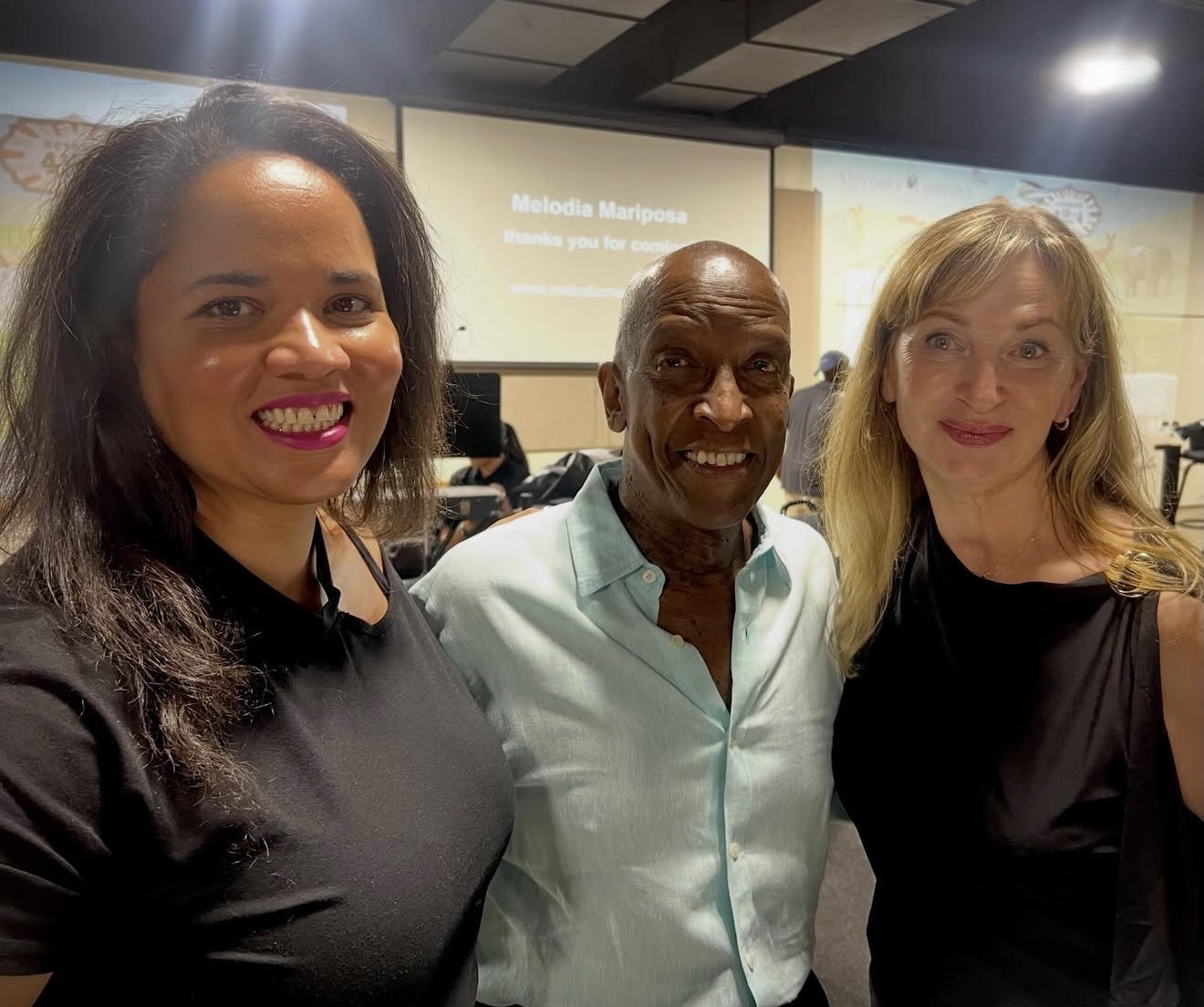 two women and a man standing in front of a projector screen that reads Melodia Mariposa 